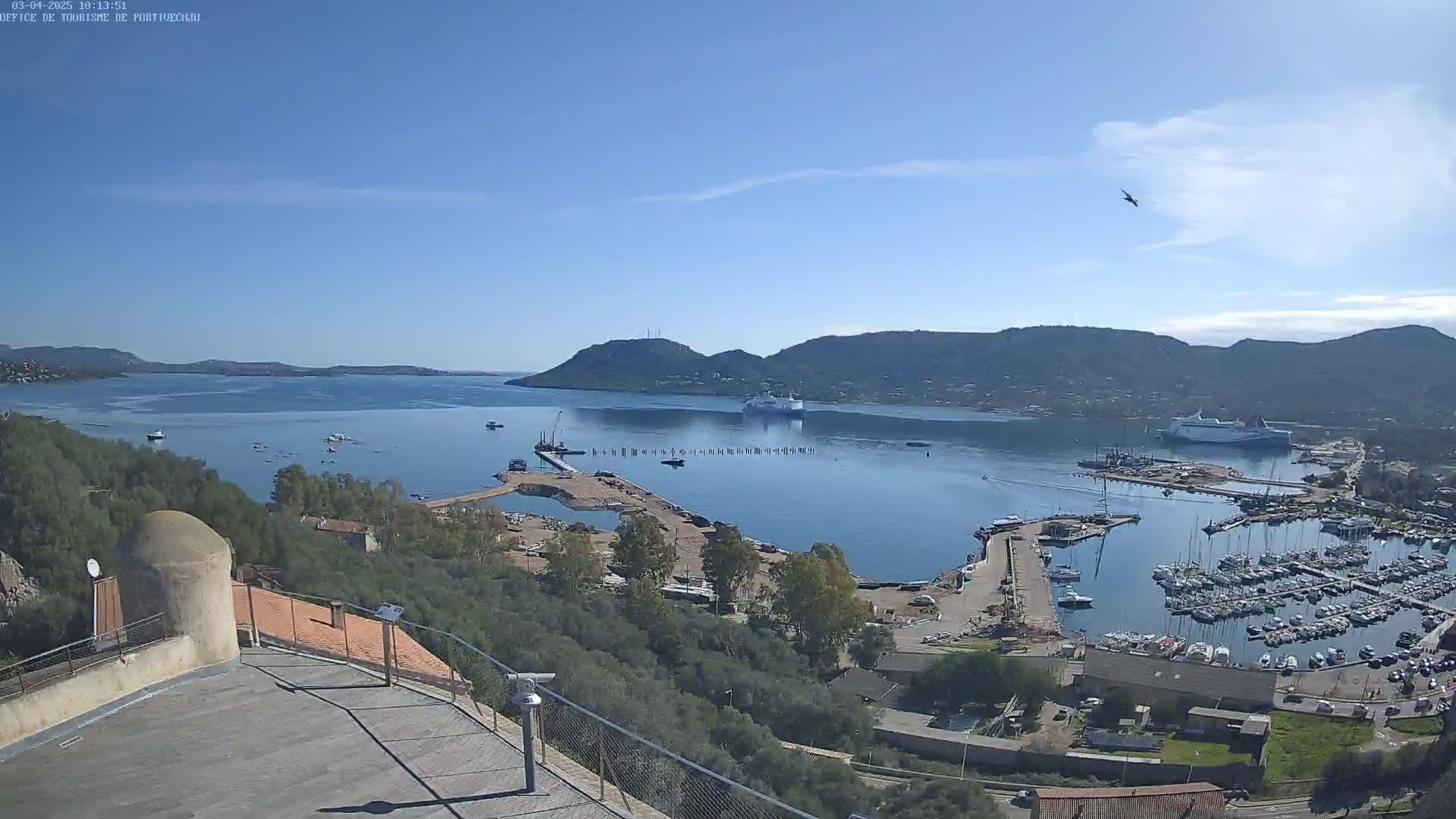 A high-angle view of a harbor filled with boats under a clear blue sky, with a large body of water and hills in the background.