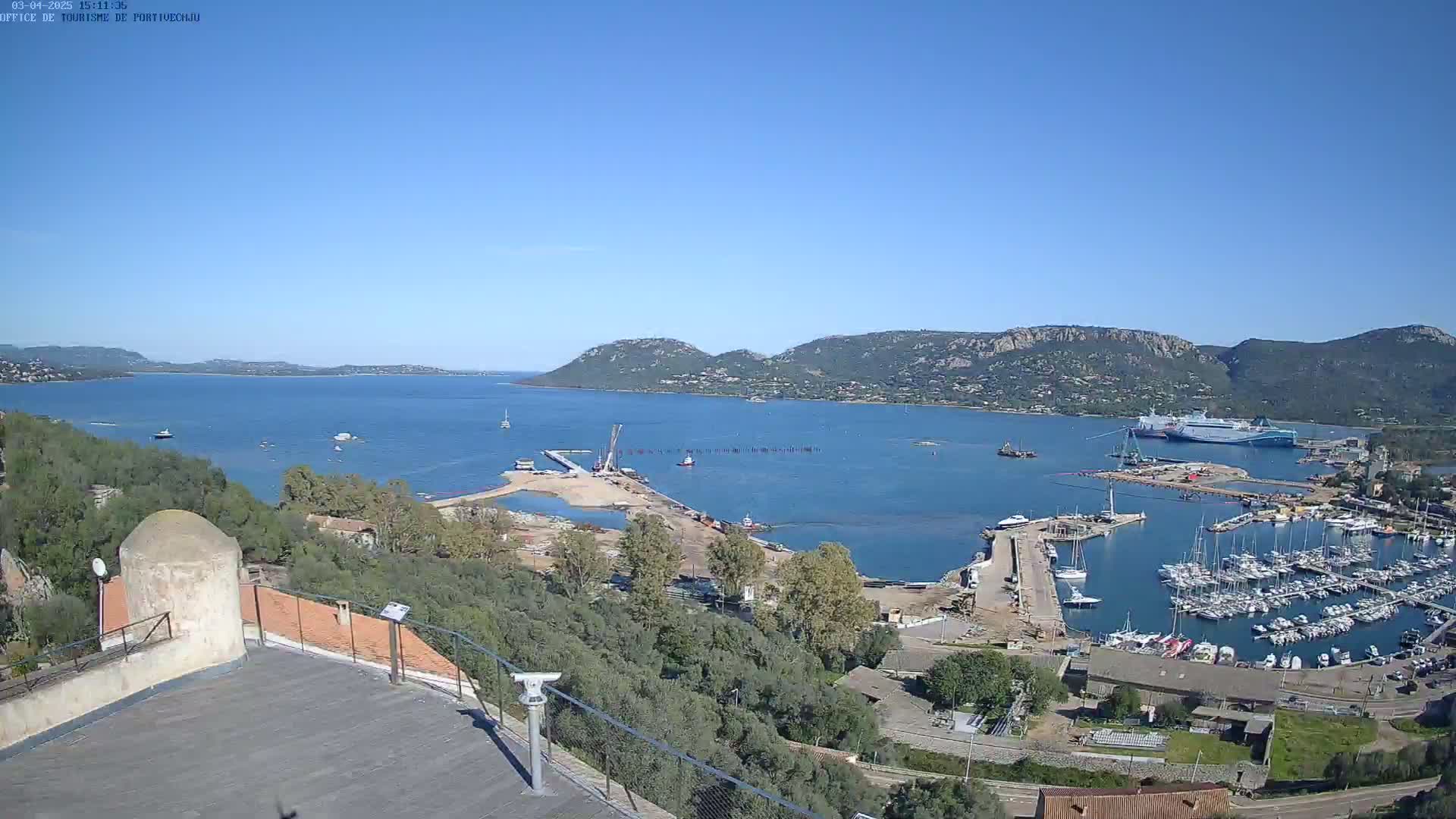 A high-angle, long shot on a clear, sunny day shows a harbor filled with boats, construction equipment, and a large ferry, situated next to a calm bay bordered by green hills.