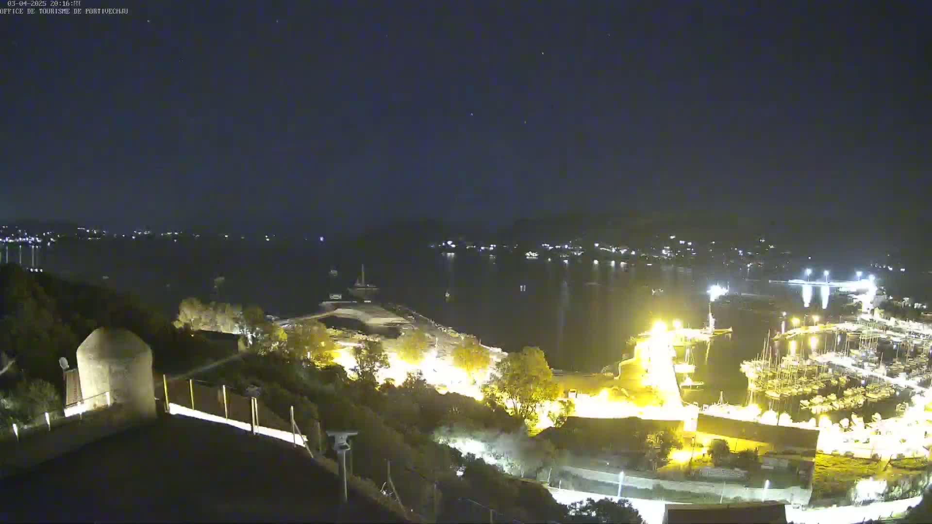 A nighttime, high-angle view shows a harbor filled with boats, illuminated buildings along the shoreline, and a dark body of water under a clear, starry sky.