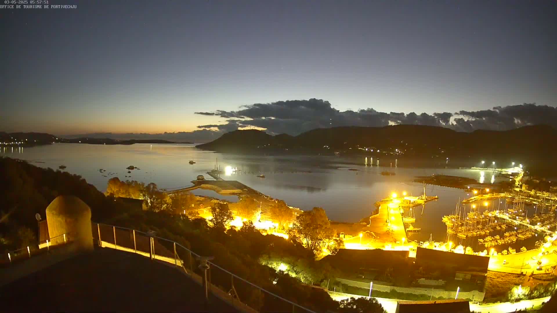 A nighttime view of a harbor filled with boats, illuminated by artificial lights, with a calm body of water reflecting the lights under a mostly clear sky with some clouds near the horizon.