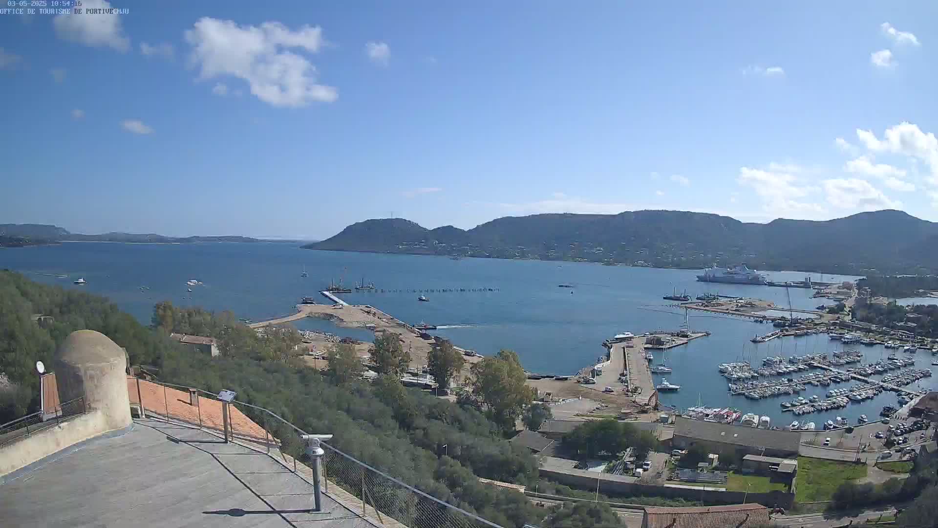 A partly sunny day overlooks a harbor filled with boats and a large ship, nestled between hills and a calm body of water.