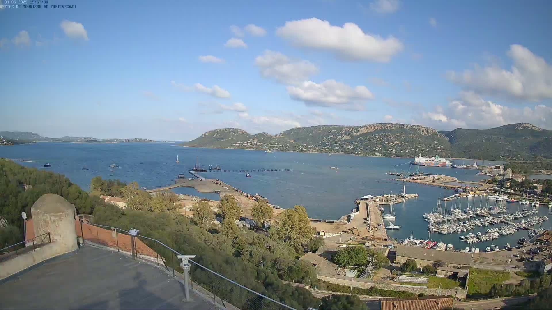 An aerial view shows a harbor filled with boats under a partly cloudy sky, with a larger body of water and distant hills in the background.