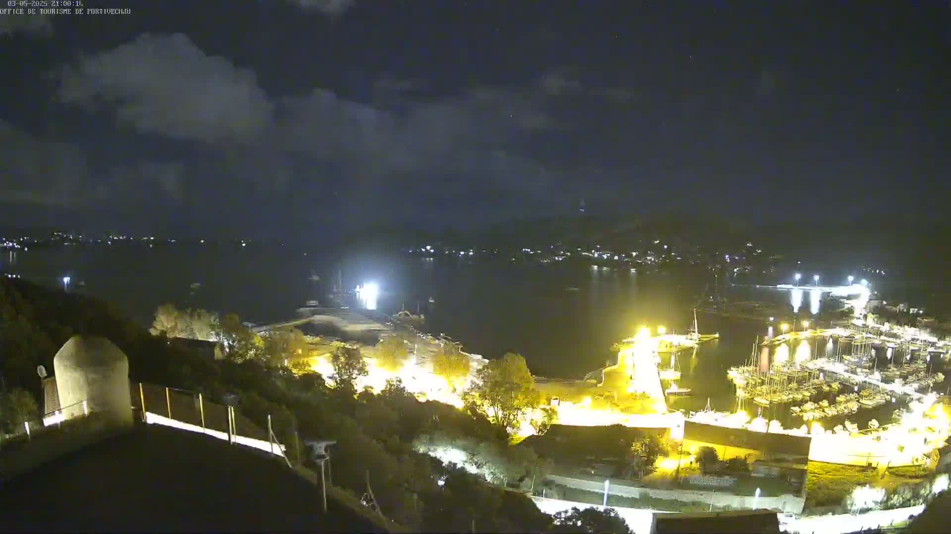 A nighttime, partly cloudy view overlooking a harbor filled with boats, lit up with artificial lights.