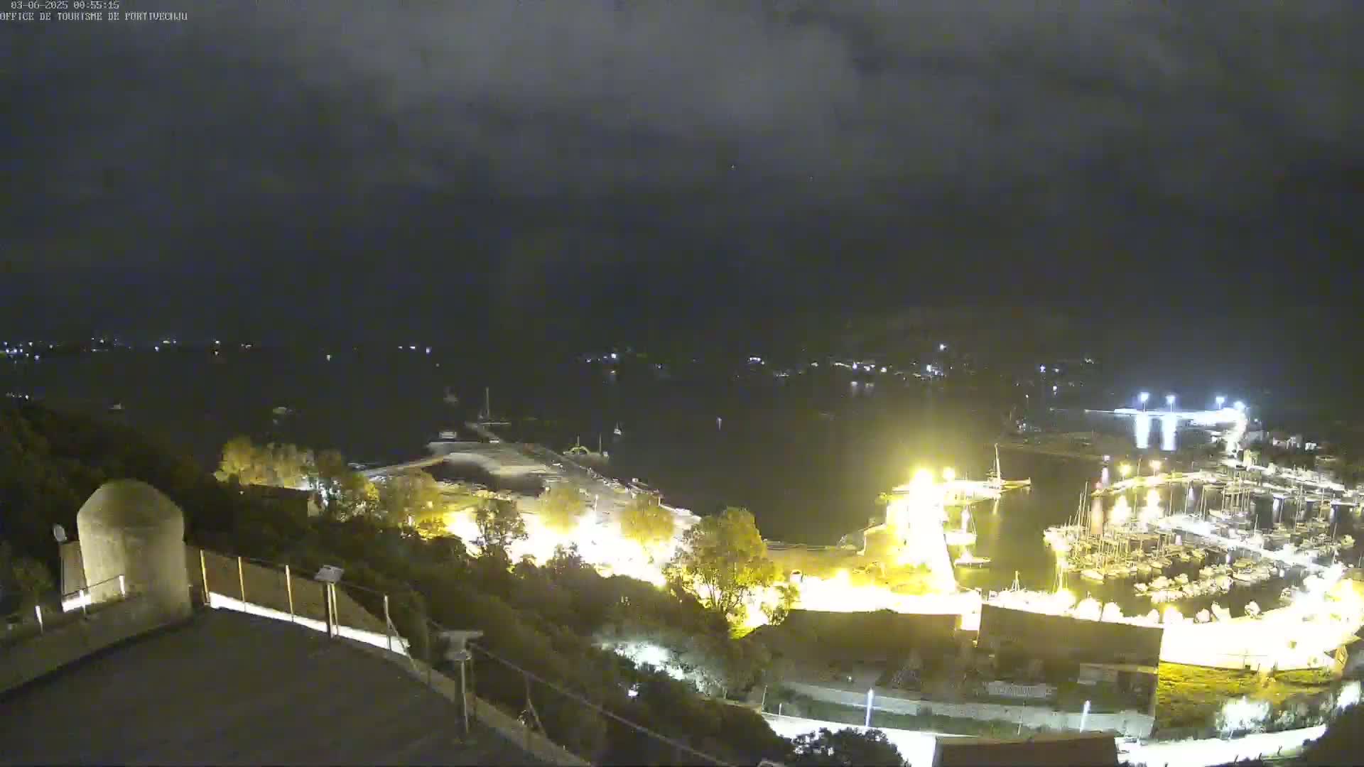 A nighttime view of a harbor filled with boats, illuminated by lights, under a partly cloudy sky.