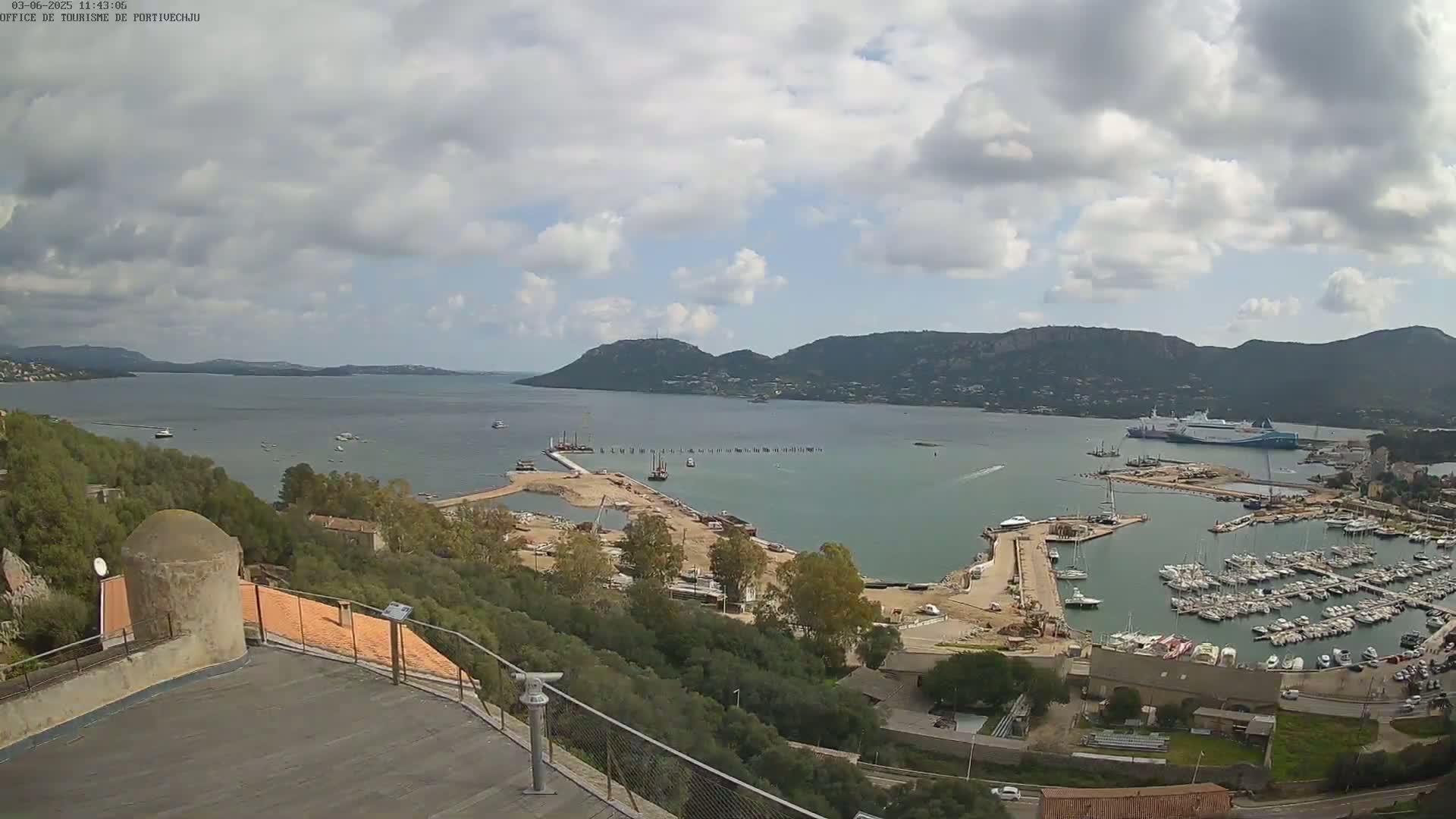 A partly cloudy day overlooks a harbor filled with numerous boats and ships, nestled between hills and a calm body of water.