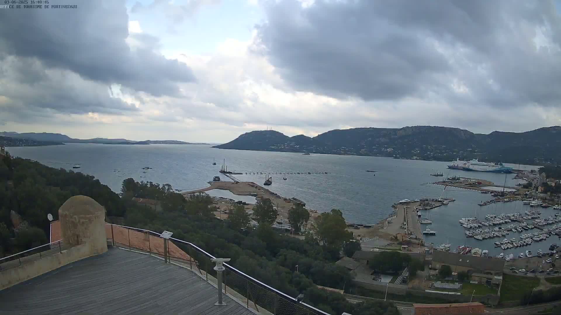 A high-angle view of a harbor filled with boats, under a cloudy sky, with hills in the background.