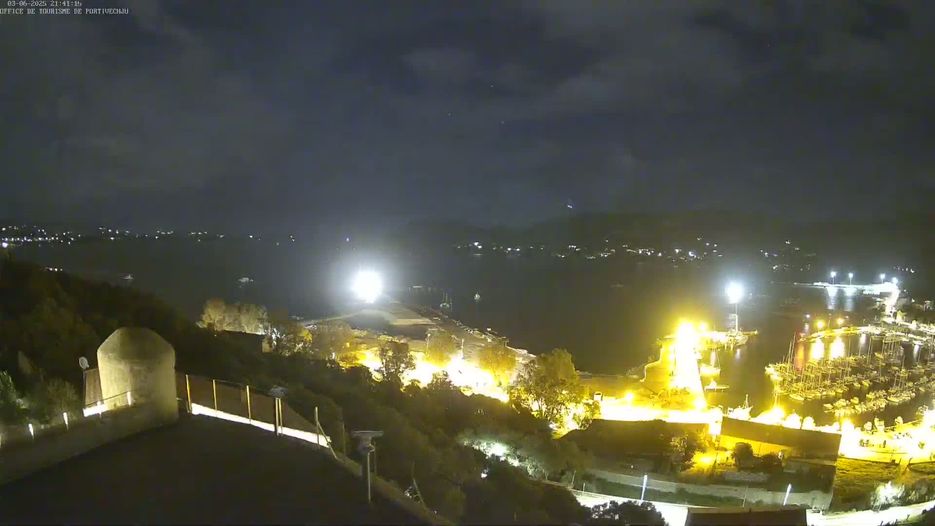 A nighttime view of a harbor filled with boats, brightly lit from shore, under a partly cloudy sky.