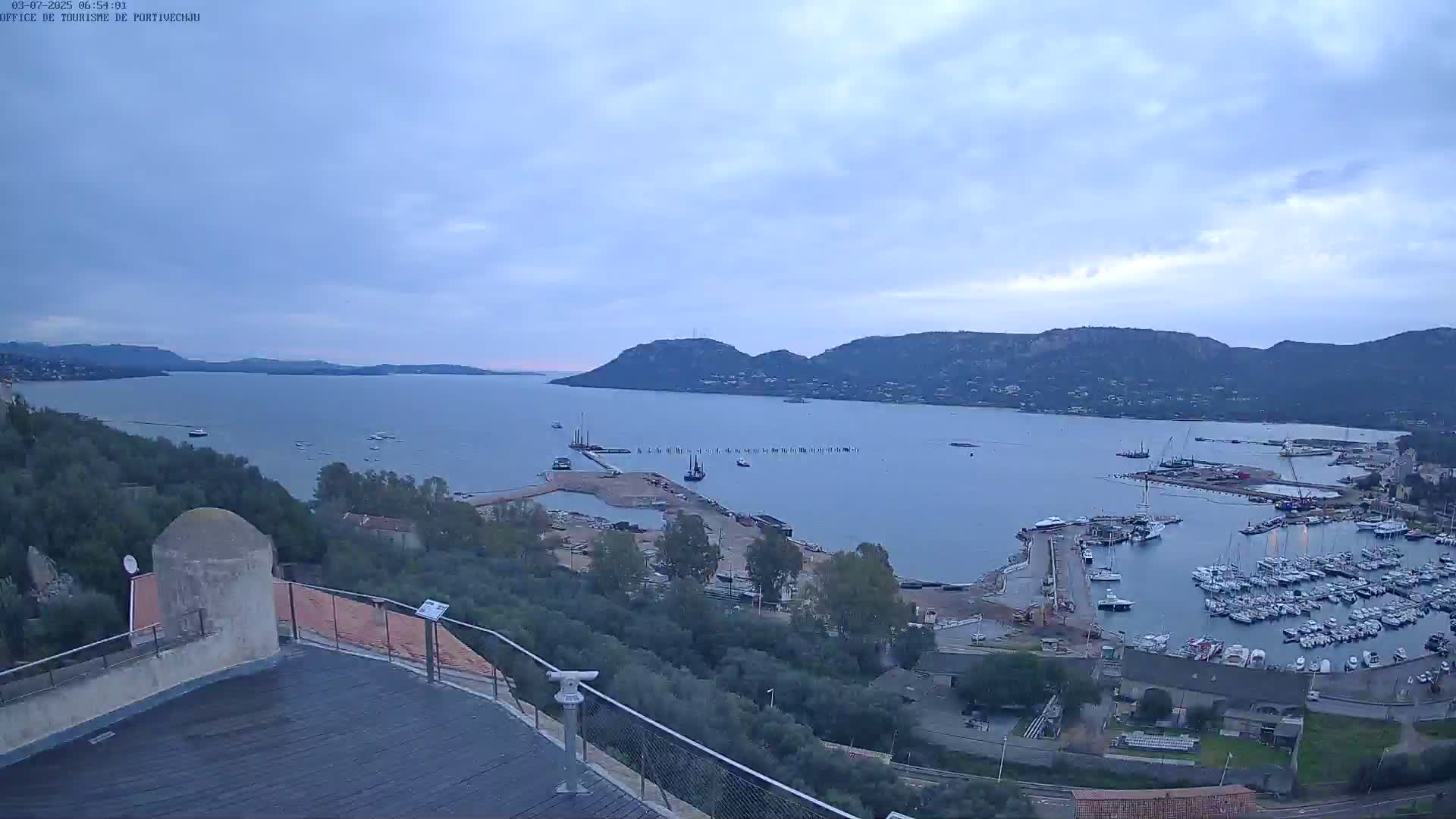 A high-angle, long shot shows a calm bay with numerous boats docked in a marina, under an overcast sky.