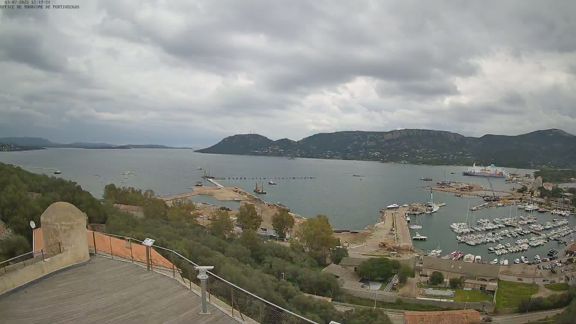 An overcast day reveals a harbor filled with numerous boats and ships, nestled beside a landmass with hills and greenery, viewed from an elevated vantage point.
