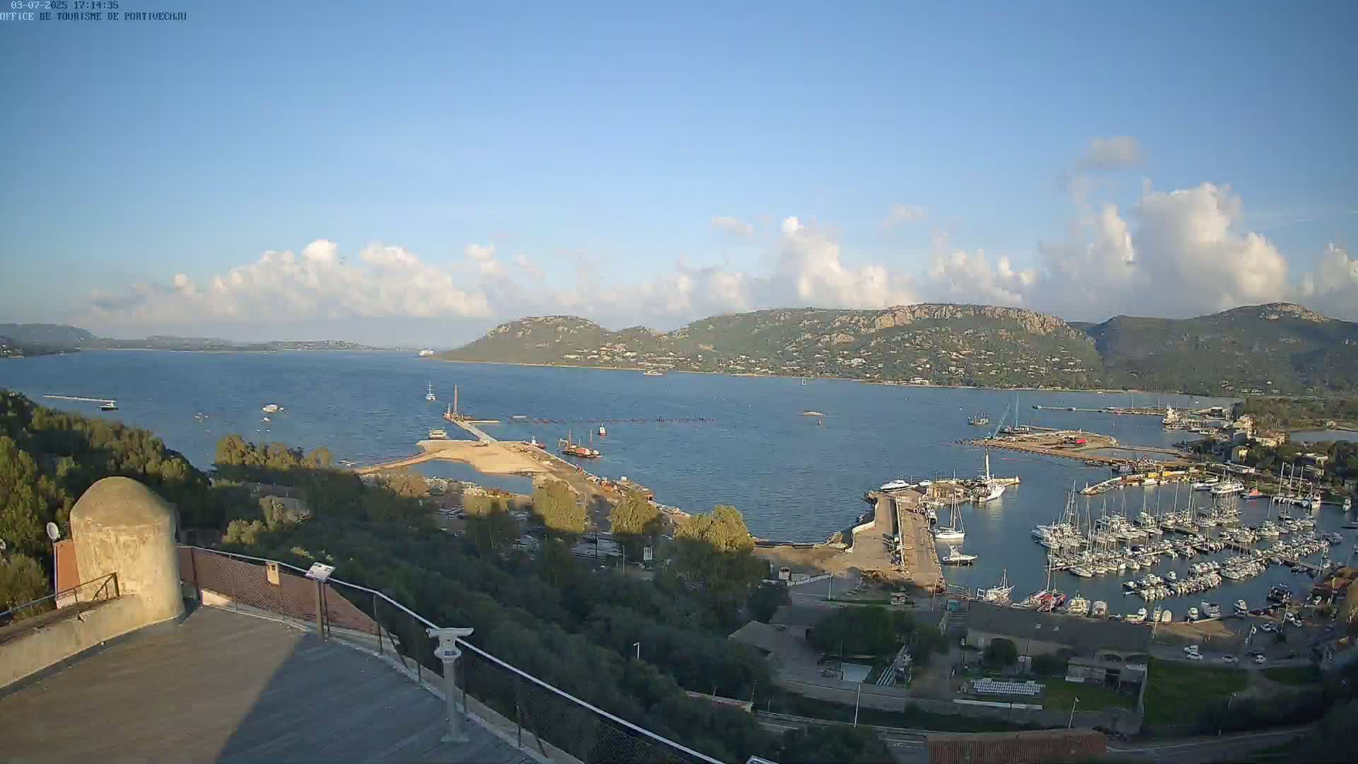 An aerial view shows a partly sunny day overlooking a harbor filled with boats, construction, and hills in the background.