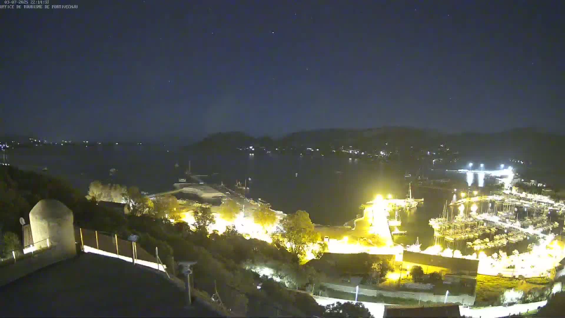 A nighttime, high-angle view of a harbor filled with boats, illuminated by artificial lights, nestled between hills under a clear, starry sky.