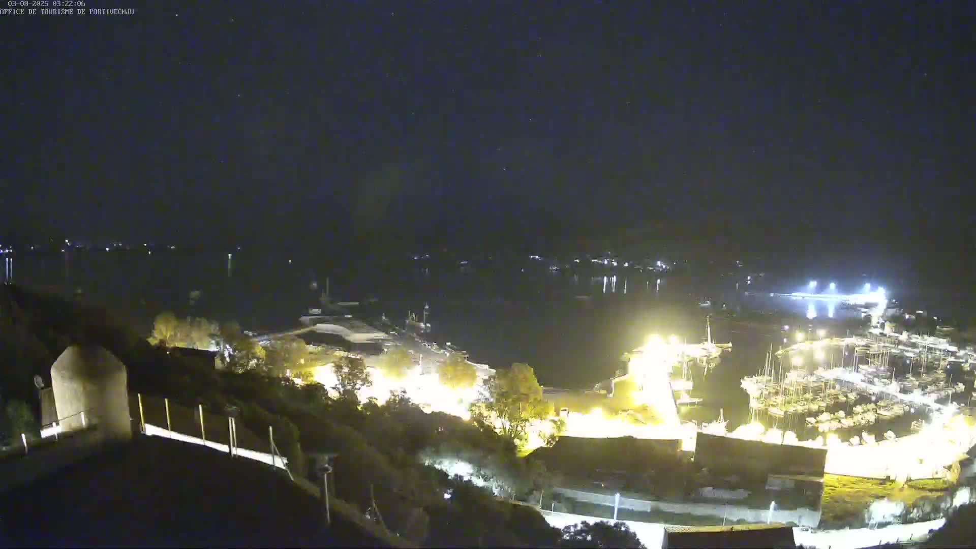 A nighttime view of a harbor filled with boats, illuminated by lights, under a clear, starry sky.