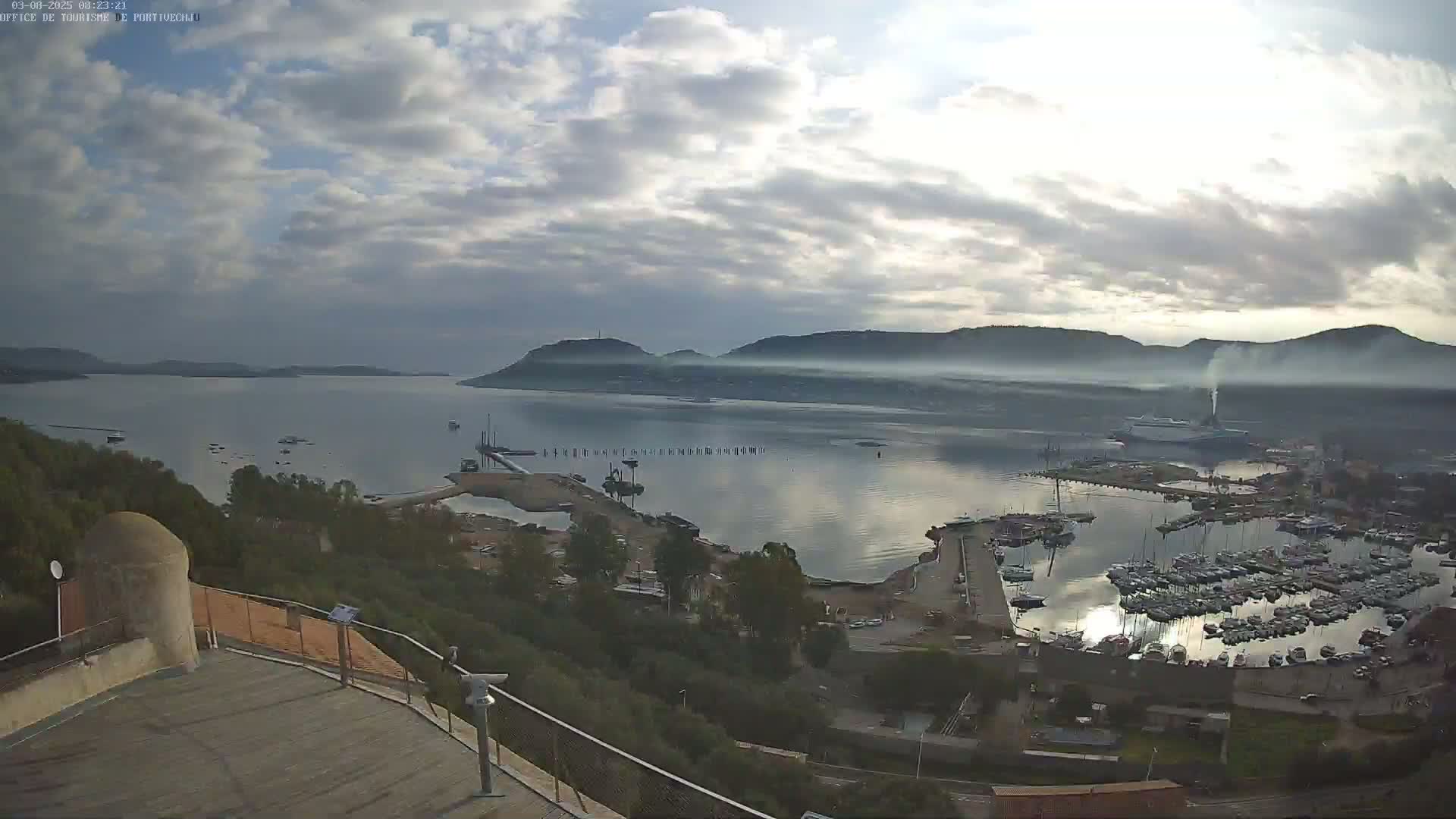An aerial view of a harbor filled with numerous boats, under a partly cloudy sky, with distant hills shrouded in fog or mist.