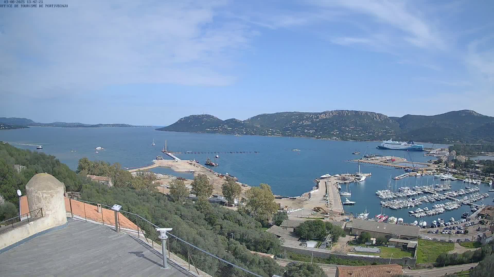 A panoramic view of a harbor filled with boats, under a mostly clear sky, with mountains in the background and construction visible near the water.