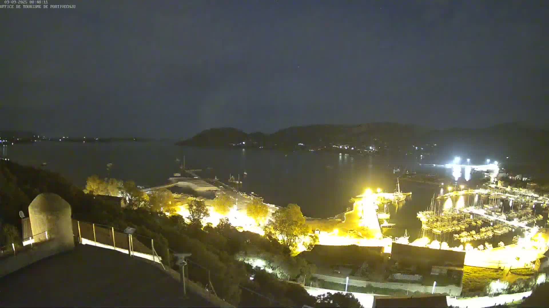 A nighttime, high-angle view shows a brightly lit harbor filled with boats, surrounded by dark hills and a calm body of water under a clear, starry sky.