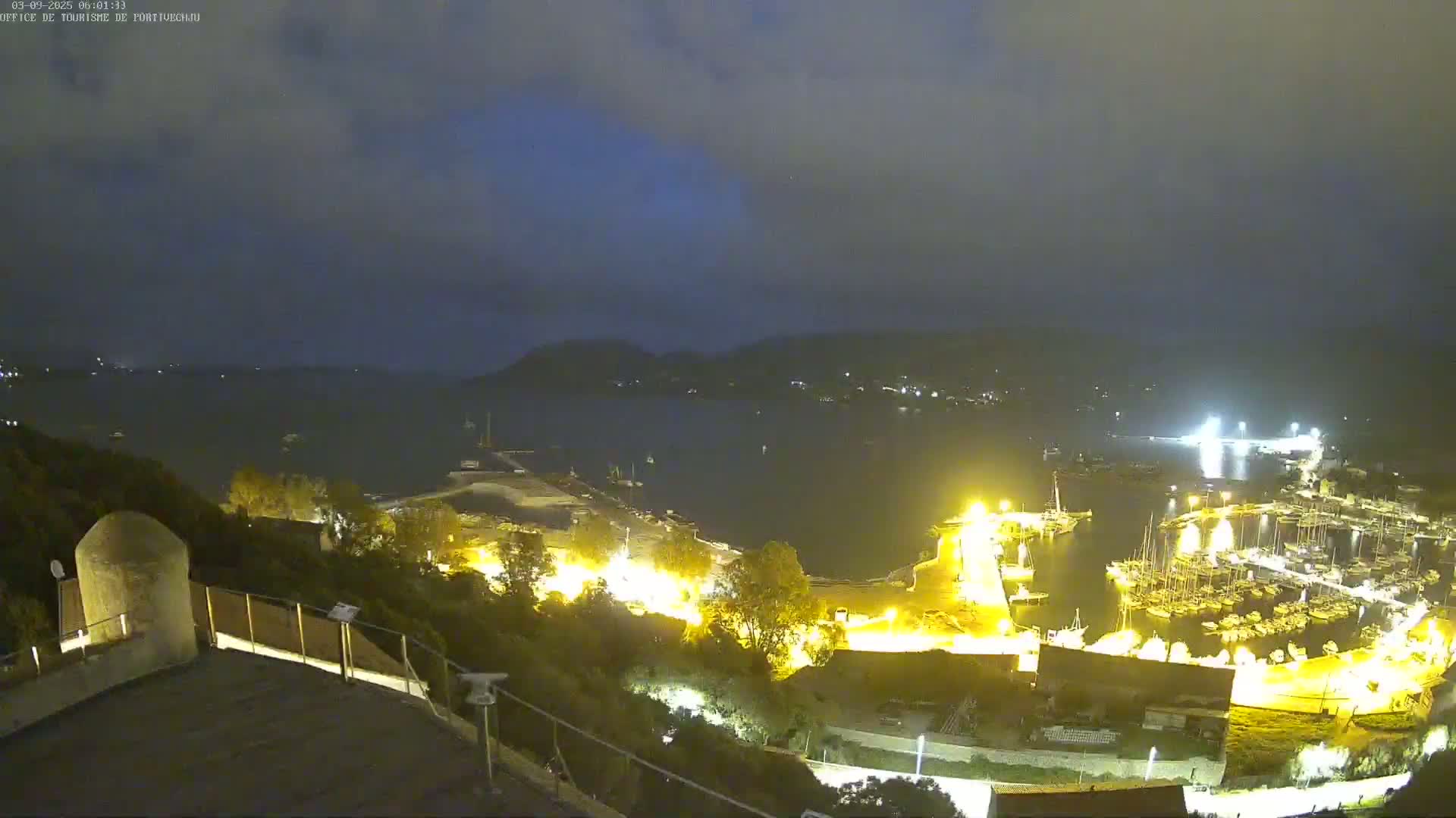 A nighttime view of a harbor filled with boats, illuminated by lights, under a partly cloudy sky.