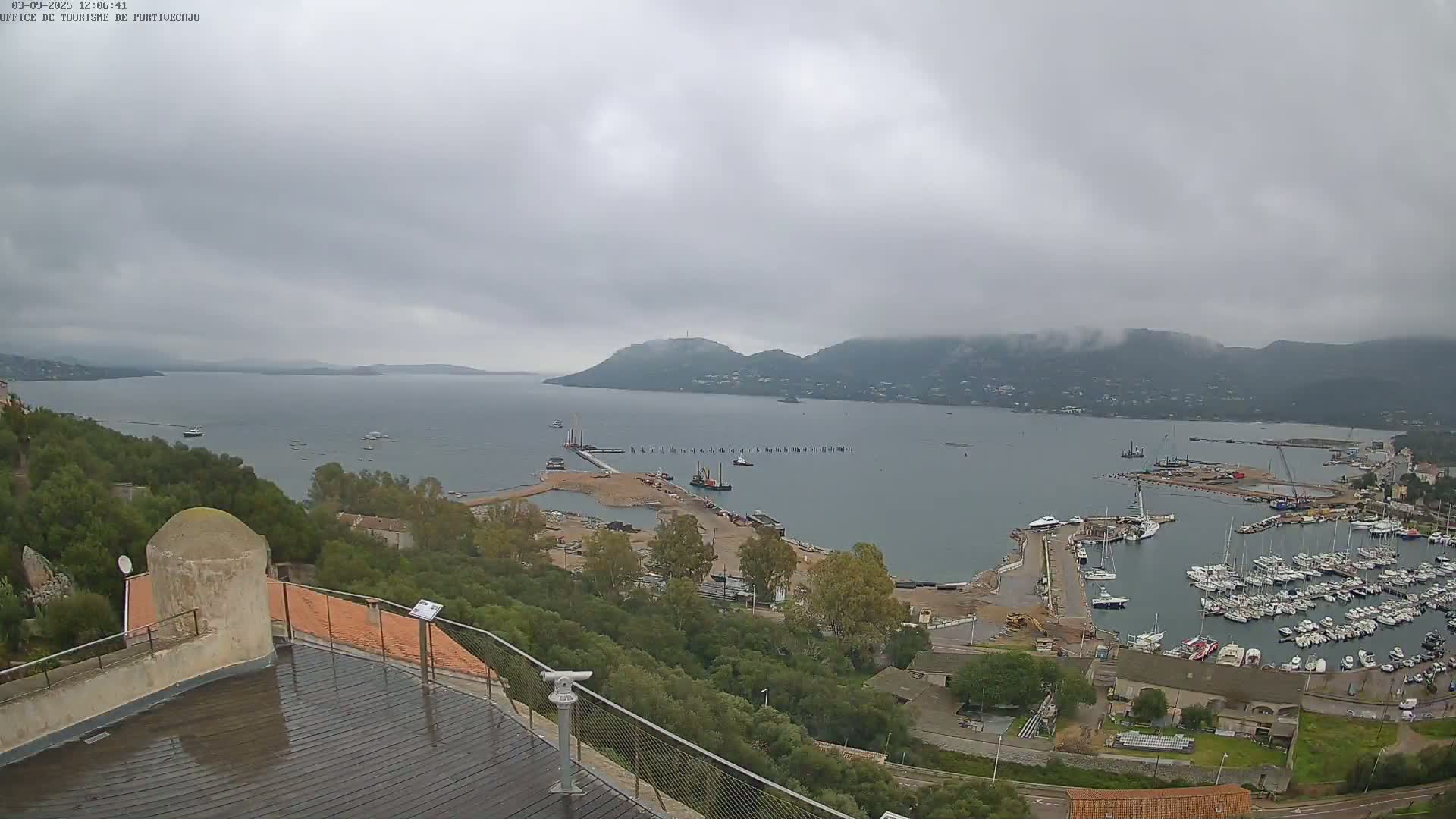 A high-angle, long shot on a cloudy day shows a harbor filled with boats, construction work on a pier, and hills shrouded in mist in the background.