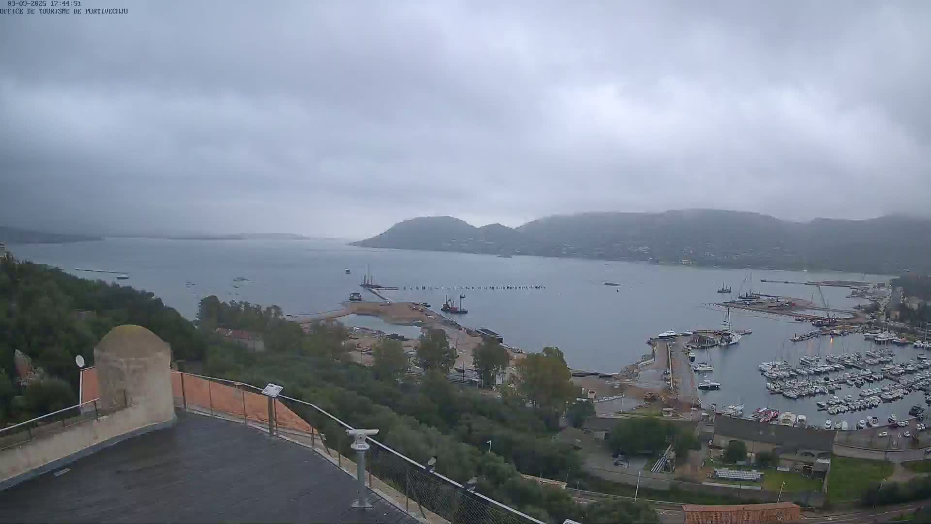 A high-angle, overcast view shows a harbor filled with boats, surrounded by land, hills, and a calm bay under a cloudy sky.