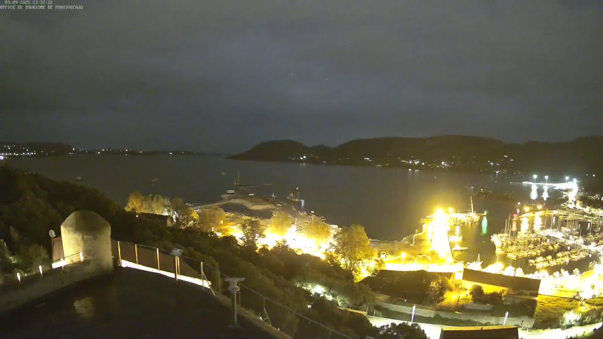 A nighttime, slightly overcast view of a harbor filled with boats, illuminated by lights, from a high vantage point overlooking a hillside.