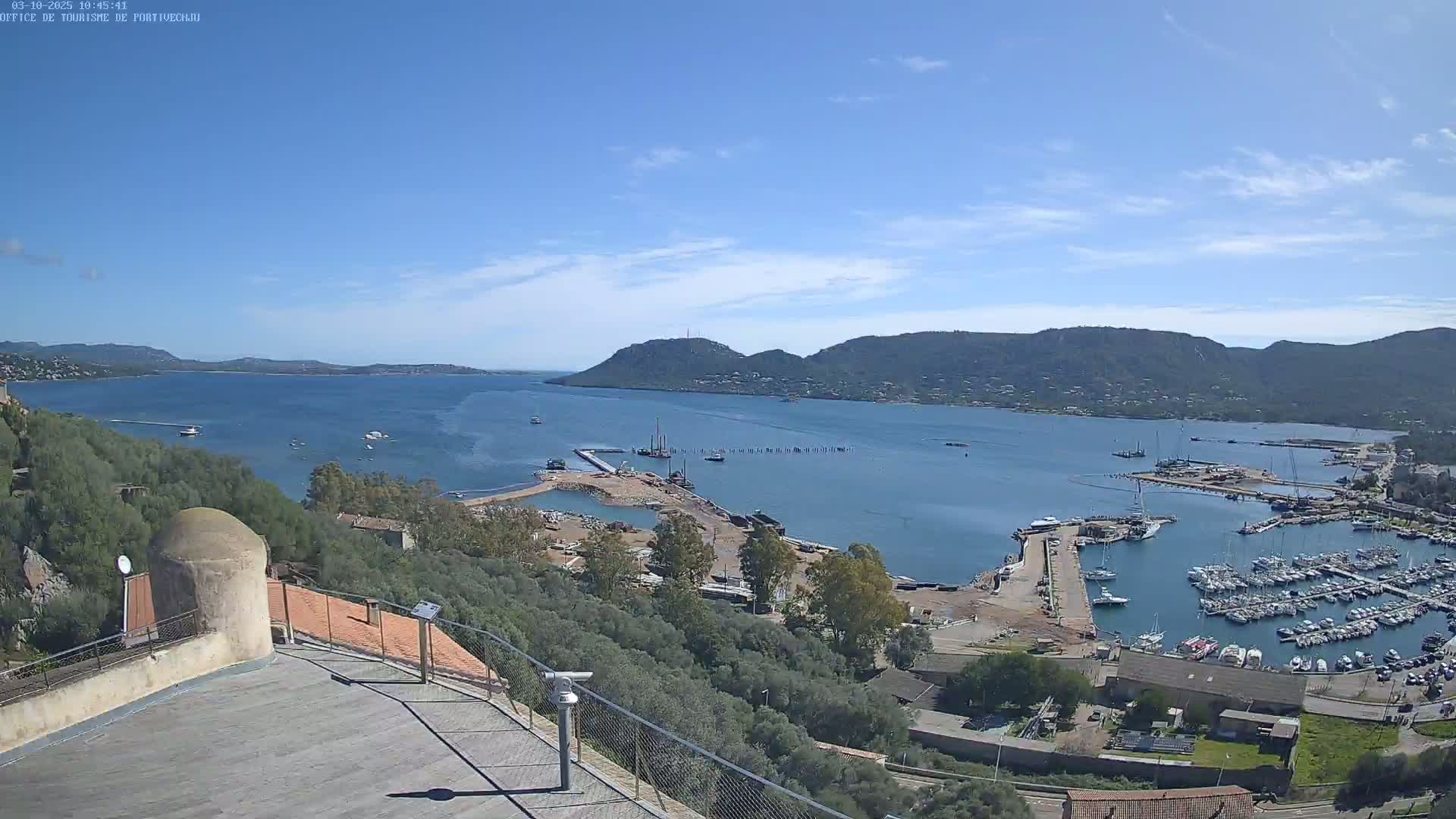 A high-angle, long shot on a clear, sunny day shows a harbor filled with boats, a construction site, and a body of water surrounded by hills.
