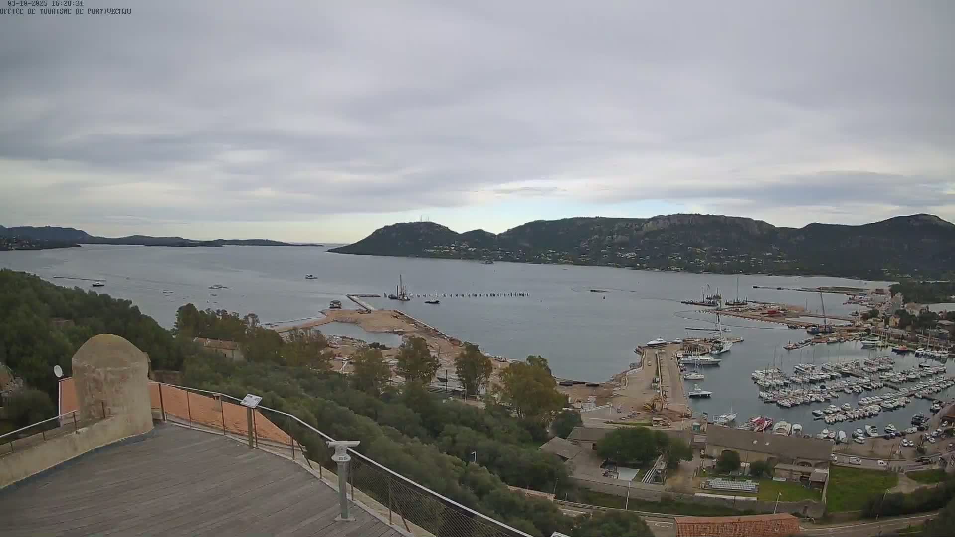 An elevated view shows a harbor filled with boats under a cloudy sky, with construction visible near the water and hills in the distance.
