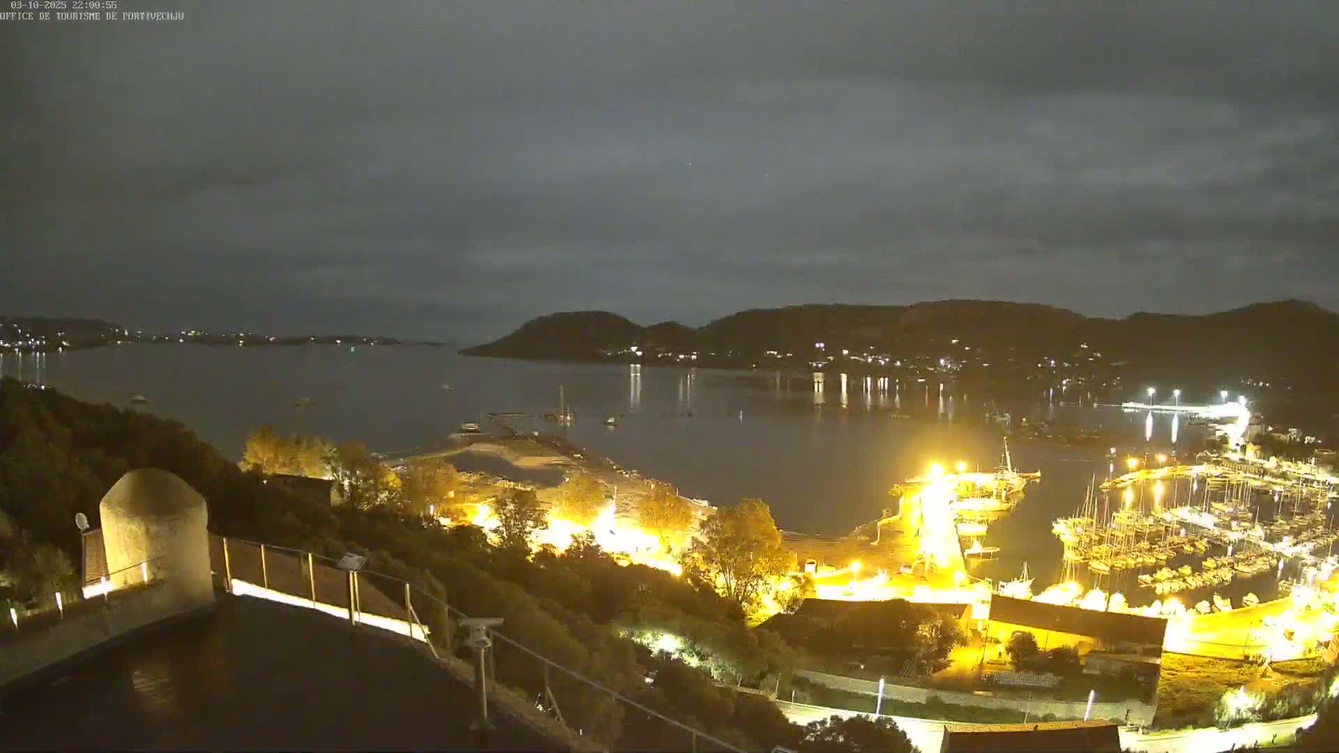 A nighttime, high-angle view shows a marina filled with boats, illuminated by lights, along a calm bay under a cloudy sky.