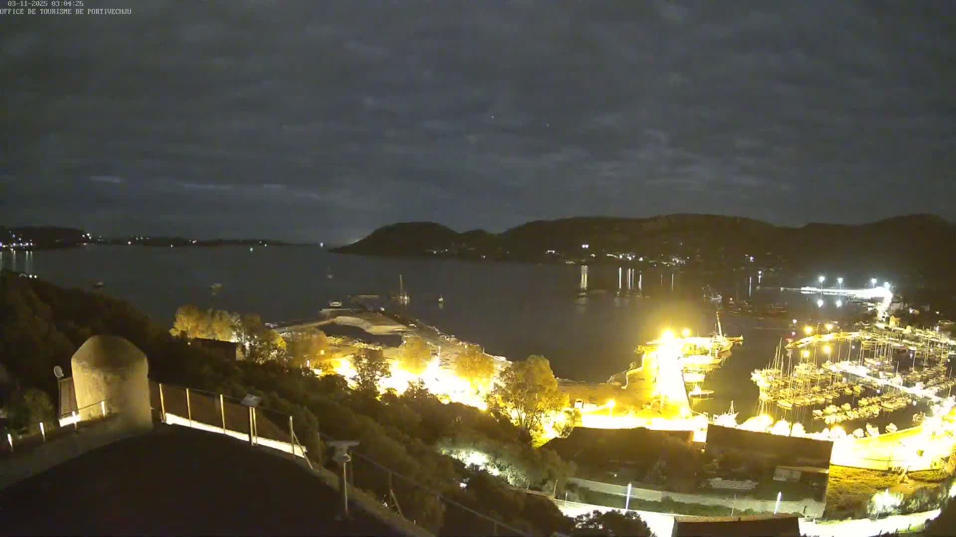 A nighttime view of a harbor filled with boats, illuminated by artificial lights, under a partly cloudy sky.