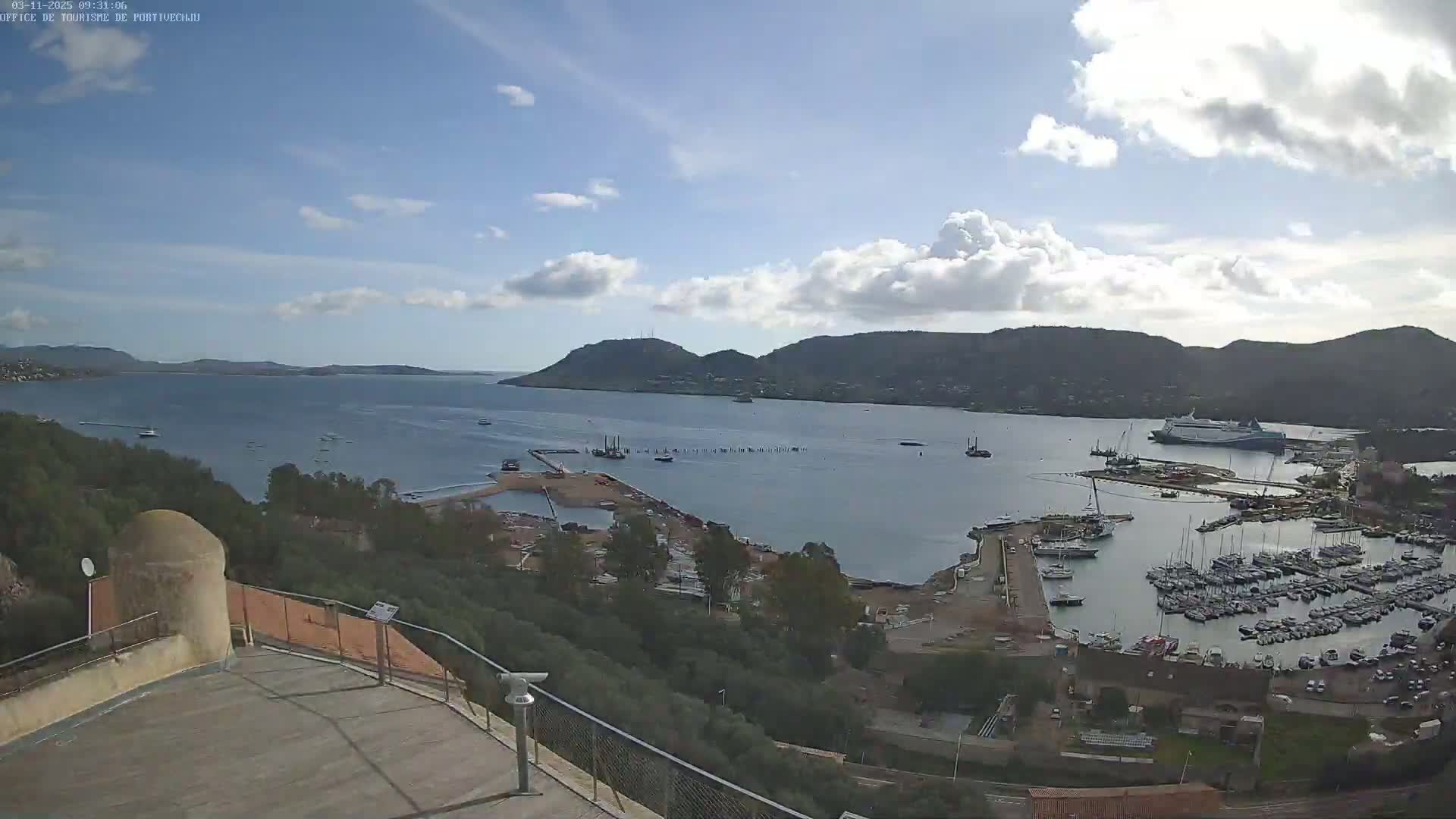 A partly cloudy day overlooks a harbor filled with numerous boats and ships, nestled between hills and a calm bay.