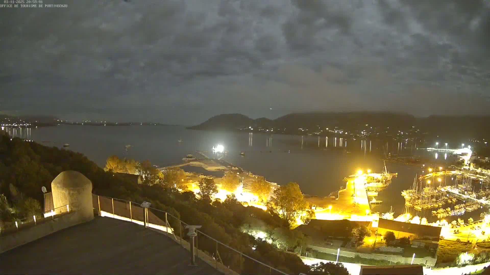 A nighttime, overcast view of a harbor filled with boats, illuminated by artificial lights, from a hilltop vantage point overlooking a calm body of water.