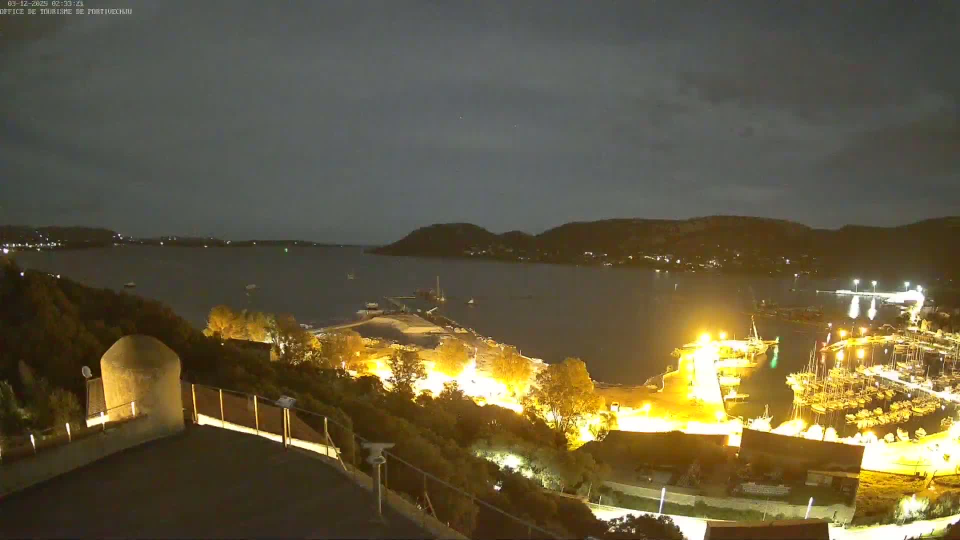 A nighttime, high-angle view of a harbor filled with boats, illuminated by artificial lights, situated on a calm body of water under a partly cloudy sky.