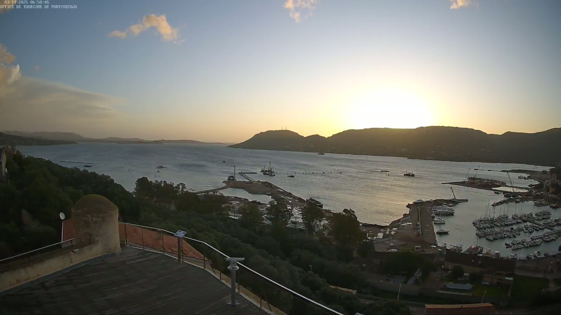 A sunset view overlooking a calm bay with a marina filled with boats, surrounded by hills and lush greenery.