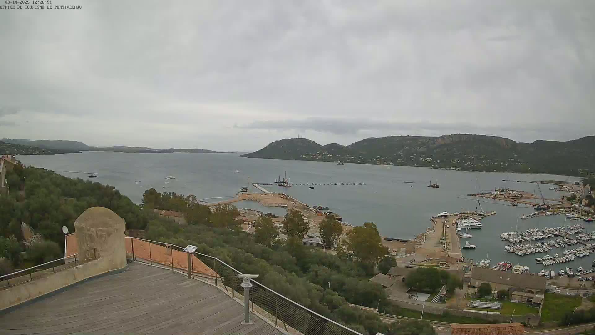 An overcast day reveals a coastal scene with a harbor filled with boats, construction underway near a marina, and a hilly landscape in the background, all viewed from an elevated position with a wooden deck and railing in the foreground.