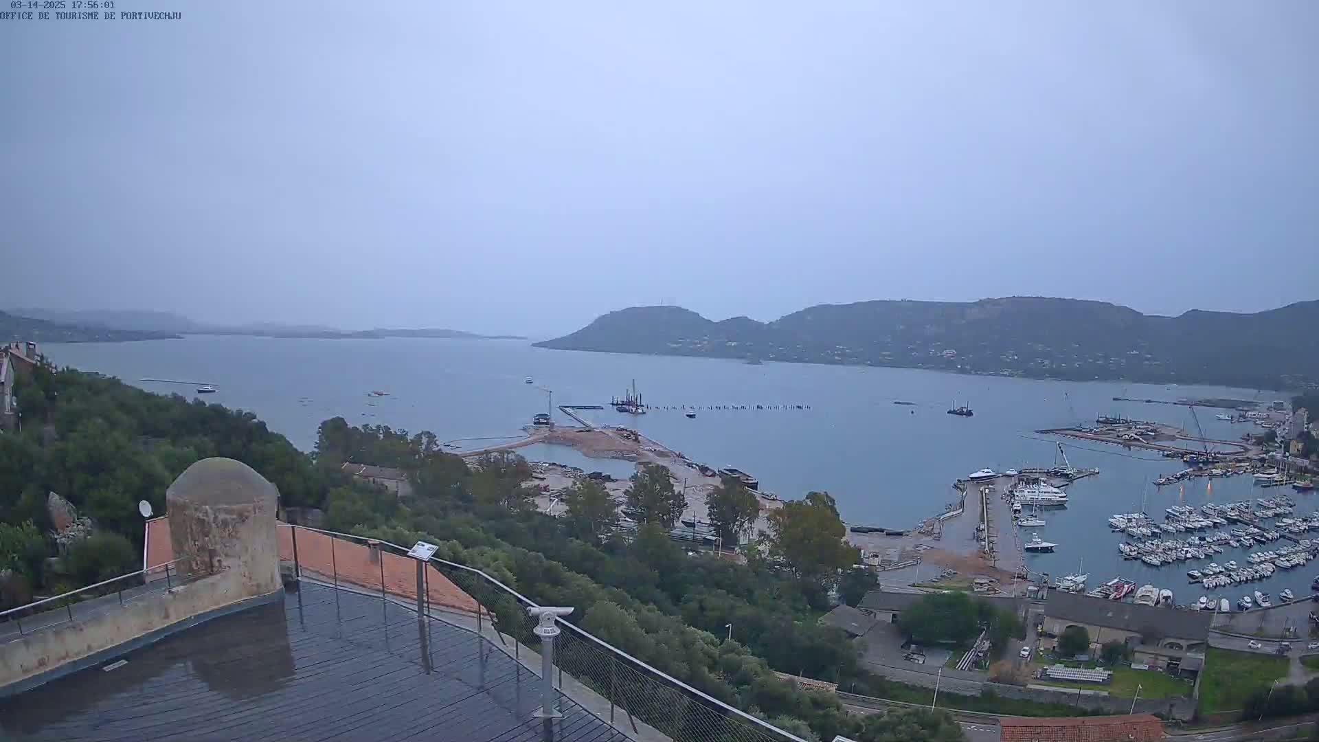 A high-angle, overcast view shows a harbor filled with boats, construction activity, and a surrounding landscape of green hills and calm water.