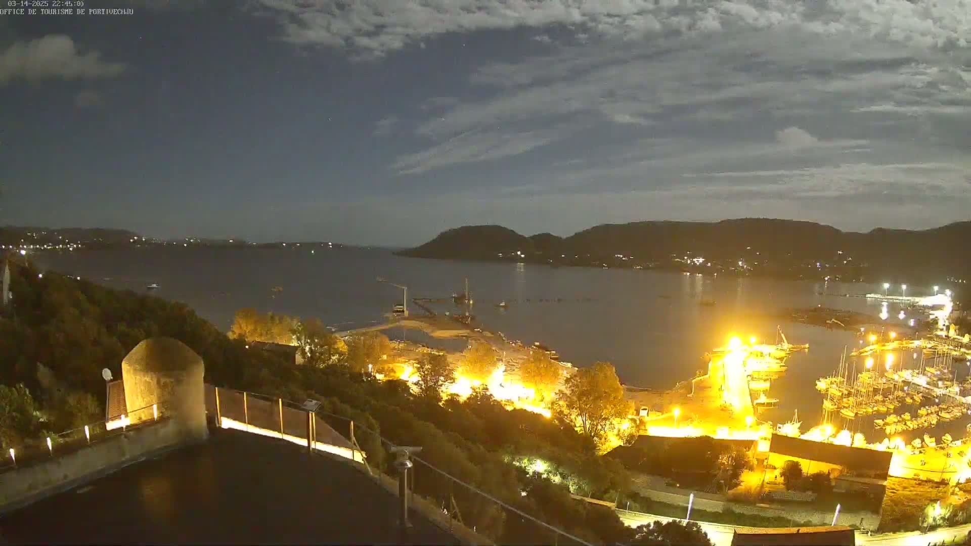 A nighttime view of a harbor with boats and buildings illuminated, under a partly cloudy sky.