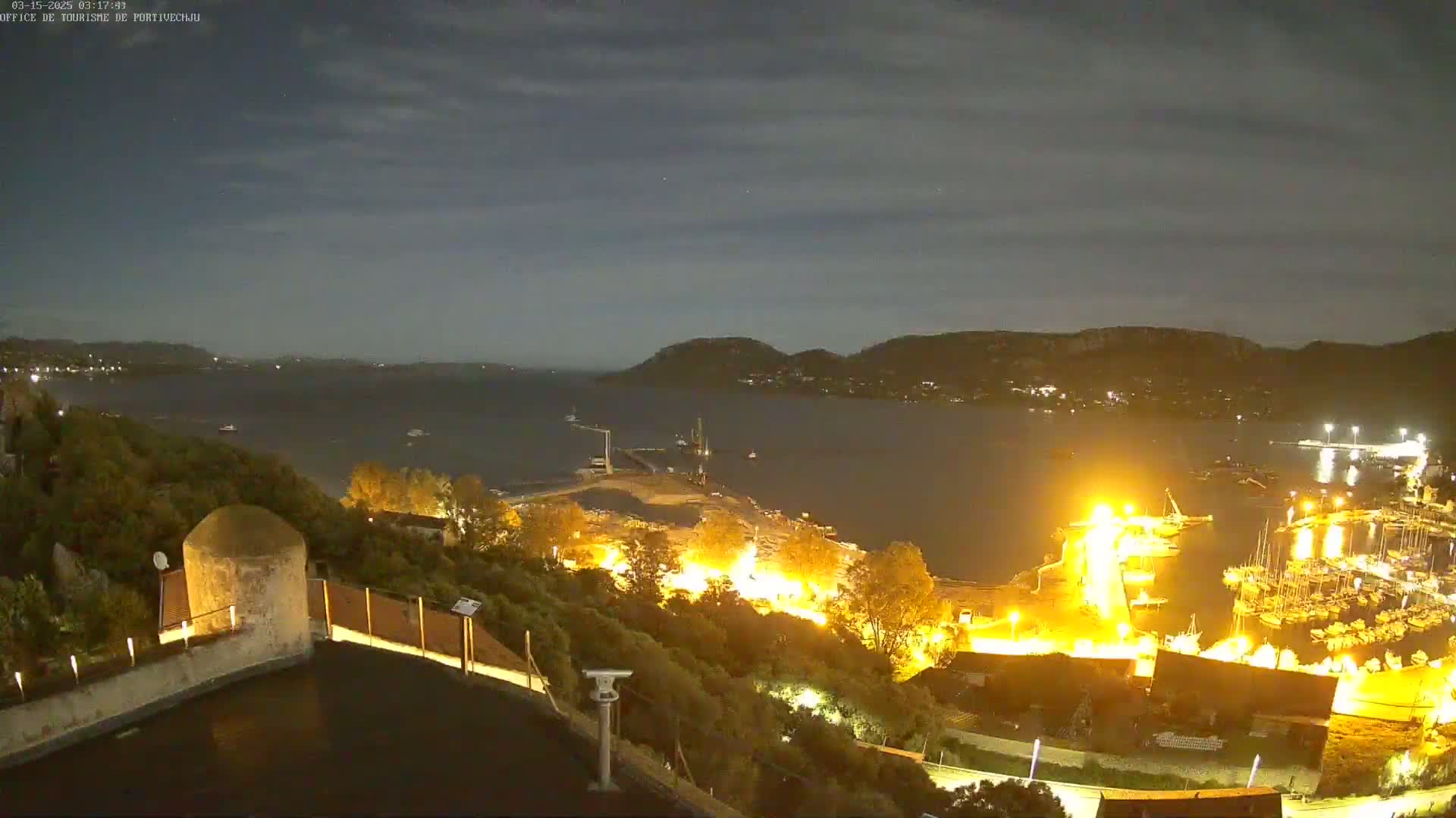 A nighttime, high-angle view shows a brightly lit harbor filled with boats, a dark body of water extending to hills under a partly cloudy sky.