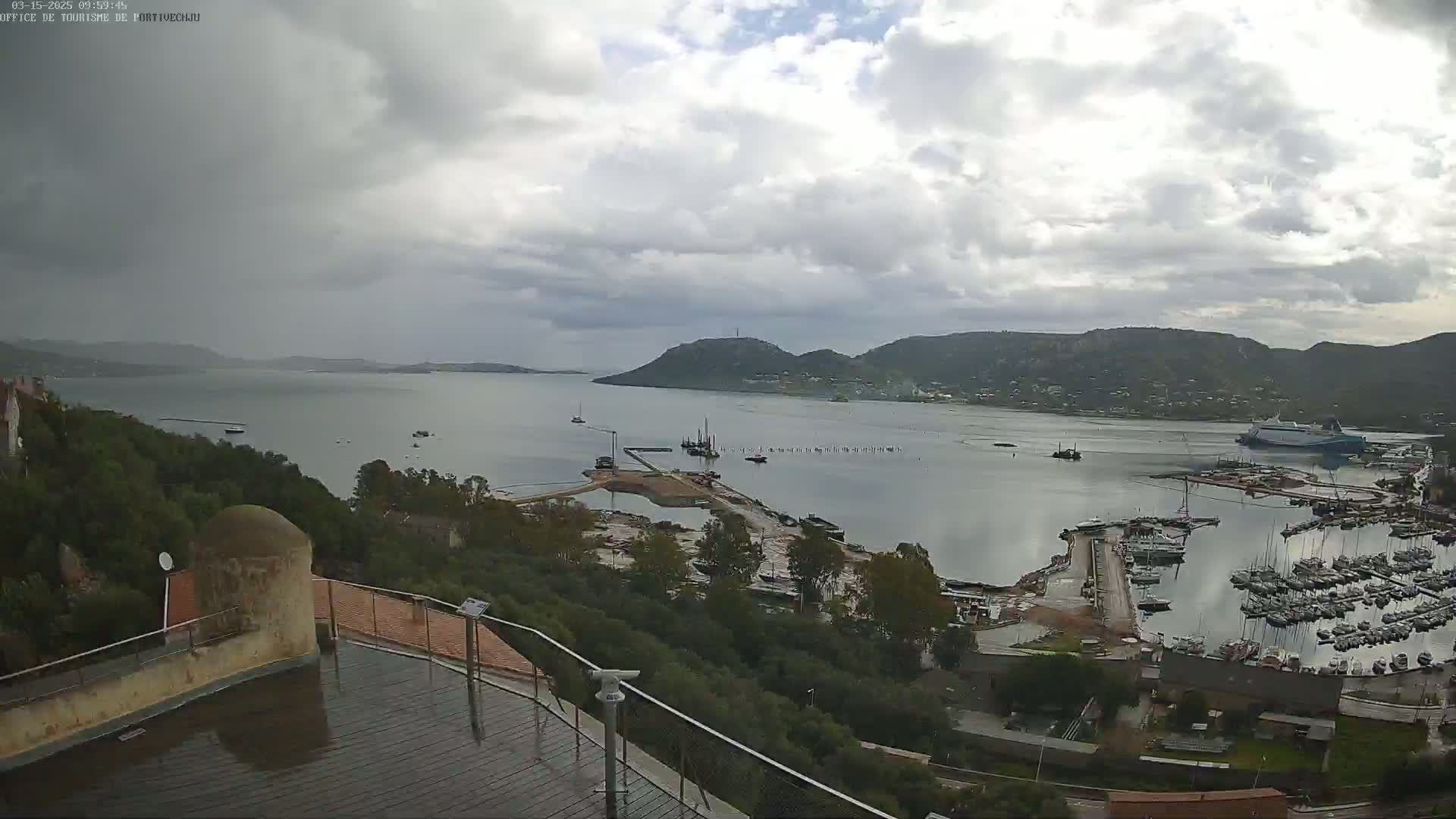 A harbor filled with boats is seen from an elevated vantage point on a cloudy day, with rain-slicked surfaces visible in the foreground.
