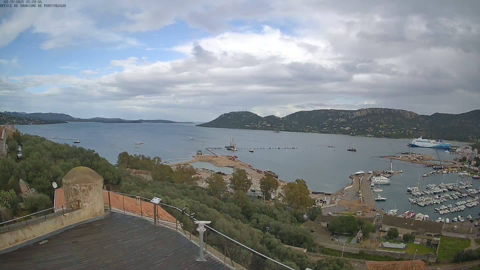 An elevated view of a harbor filled with boats, under a partly cloudy sky, shows ongoing construction work near a large body of water surrounded by hills.