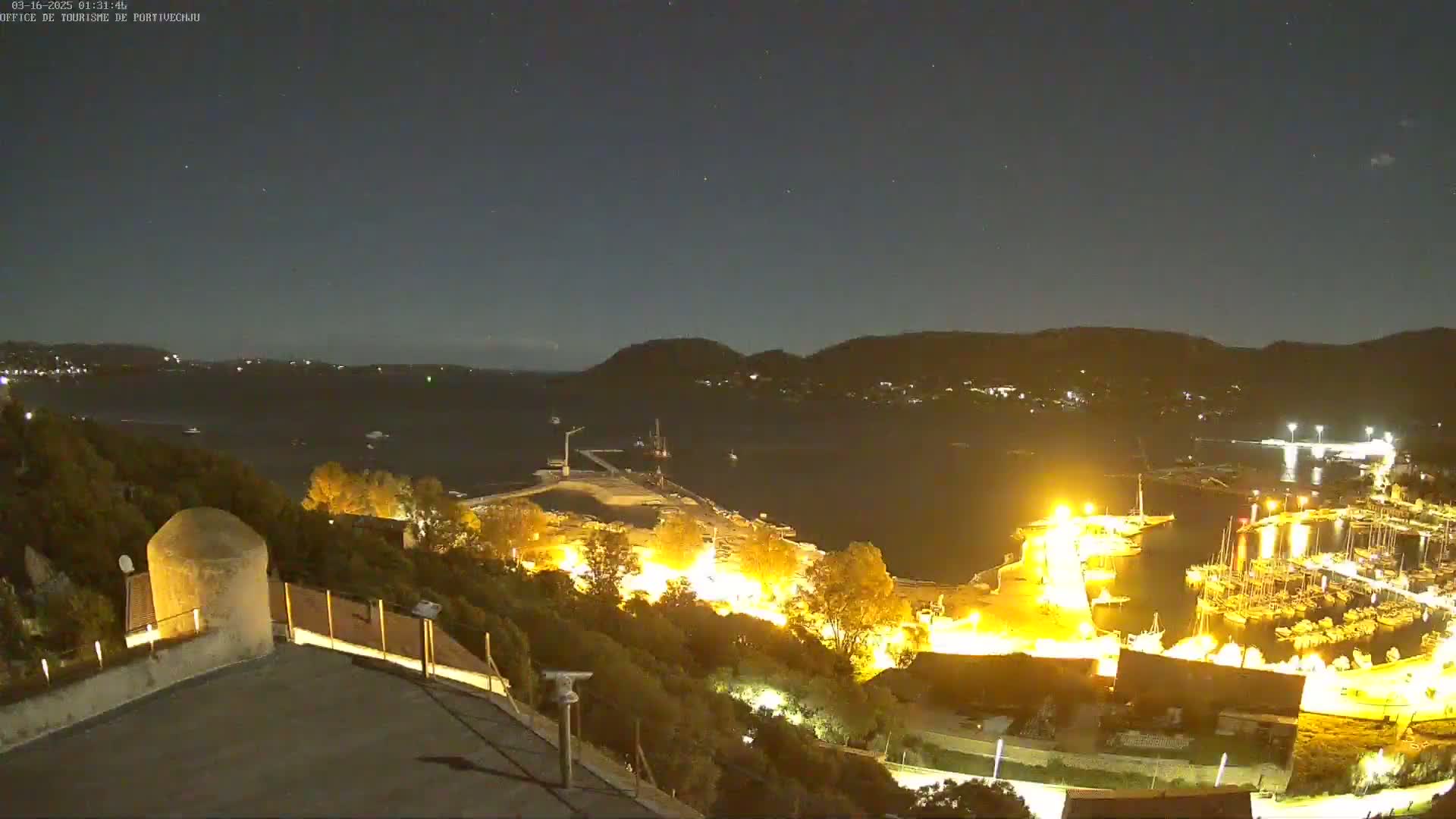 A nighttime, high-angle view of a harbor filled with boats, illuminated by artificial lights, against a backdrop of dark hills under a clear, starry sky.