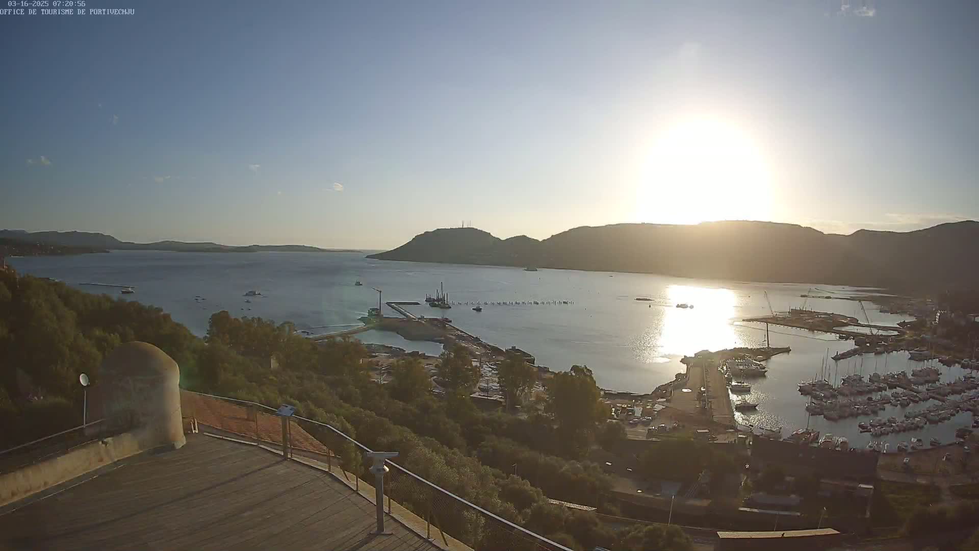 A high-angle, sunny view of a harbor filled with boats, surrounded by hills and lush green vegetation.