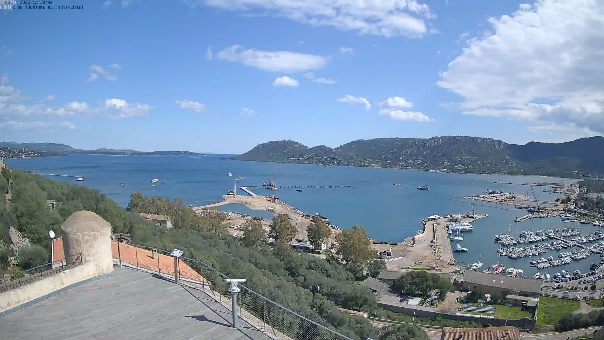 A mostly sunny day overlooking a harbor filled with boats and construction equipment, with mountains visible in the distance.
