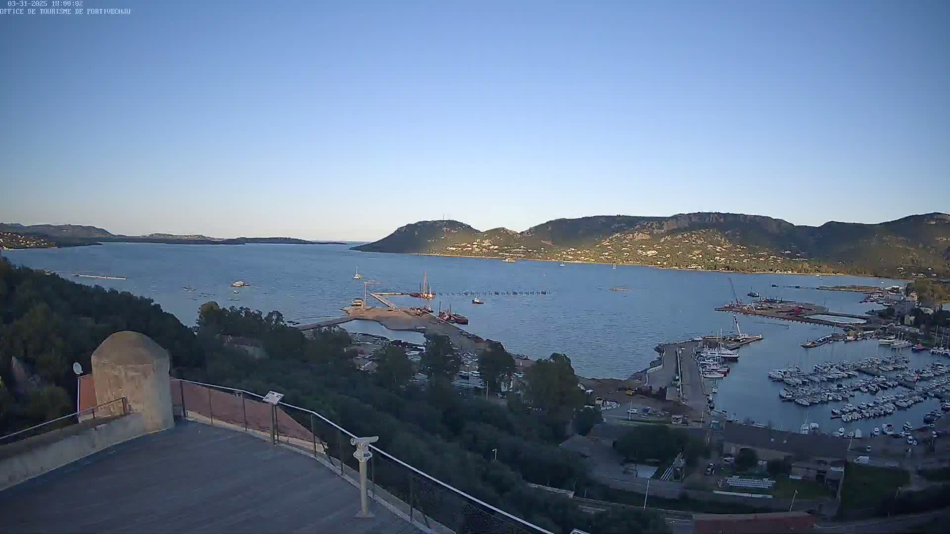 A clear, sunny day overlooks a harbor filled with boats, a small town, and a large body of water surrounded by hills.