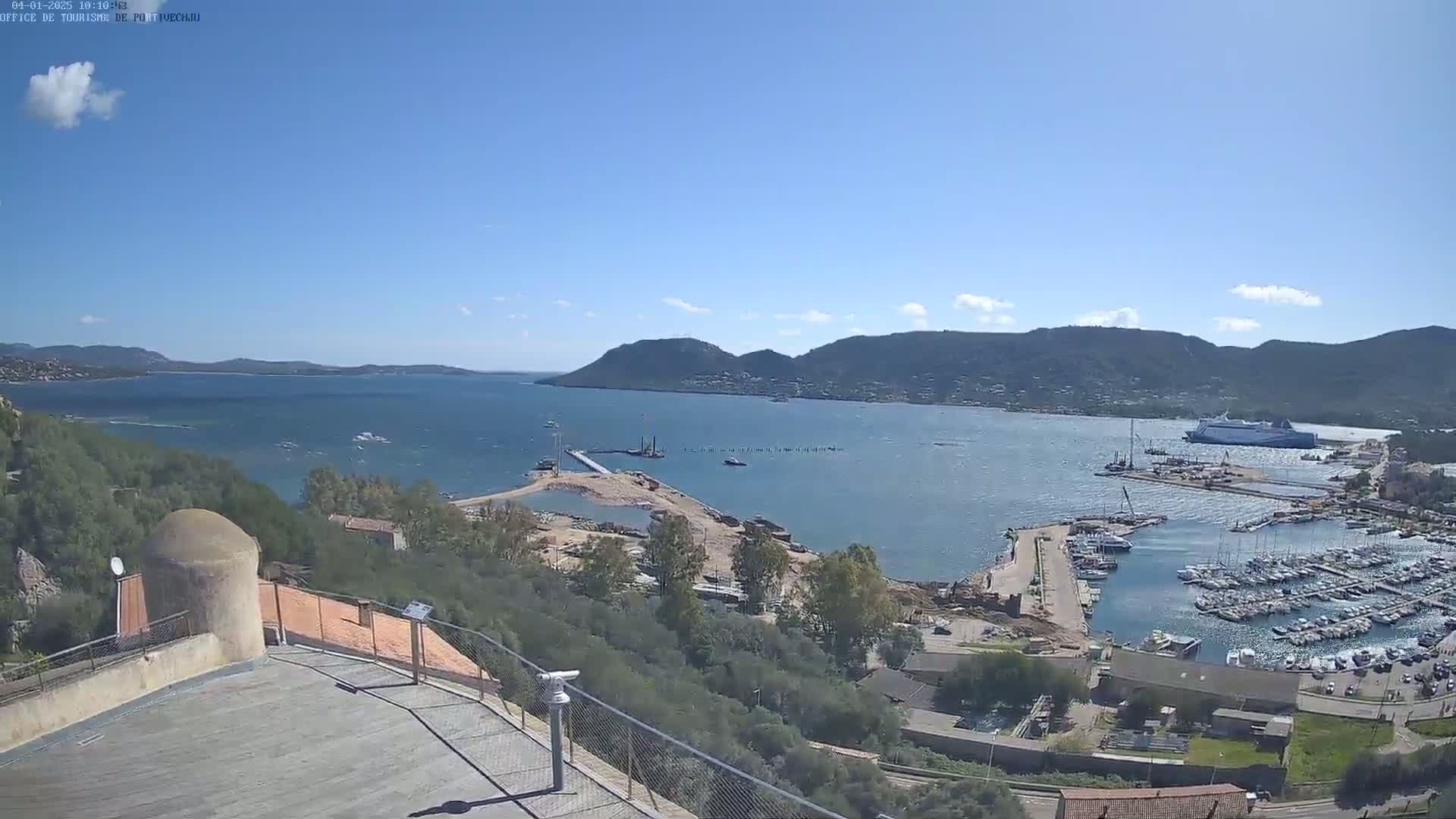 An elevated view of a harbor filled with boats, under a mostly clear blue sky, shows construction activity near a landmass extending into the water.