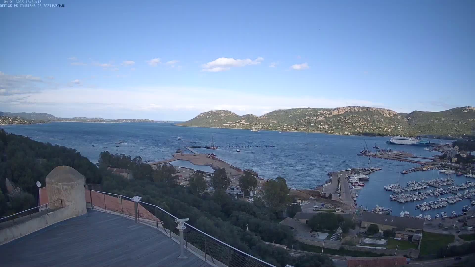 An elevated view of a harbor filled with boats, under a clear blue sky, shows a large body of water surrounded by hills and greenery.