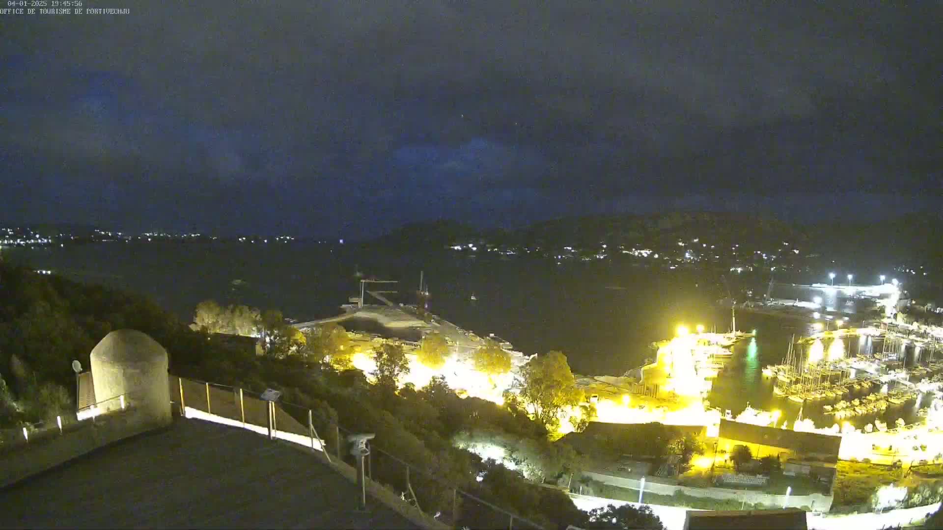 A nighttime, slightly cloudy view overlooking a harbor filled with boats, illuminated buildings, and a small structure on a hill.