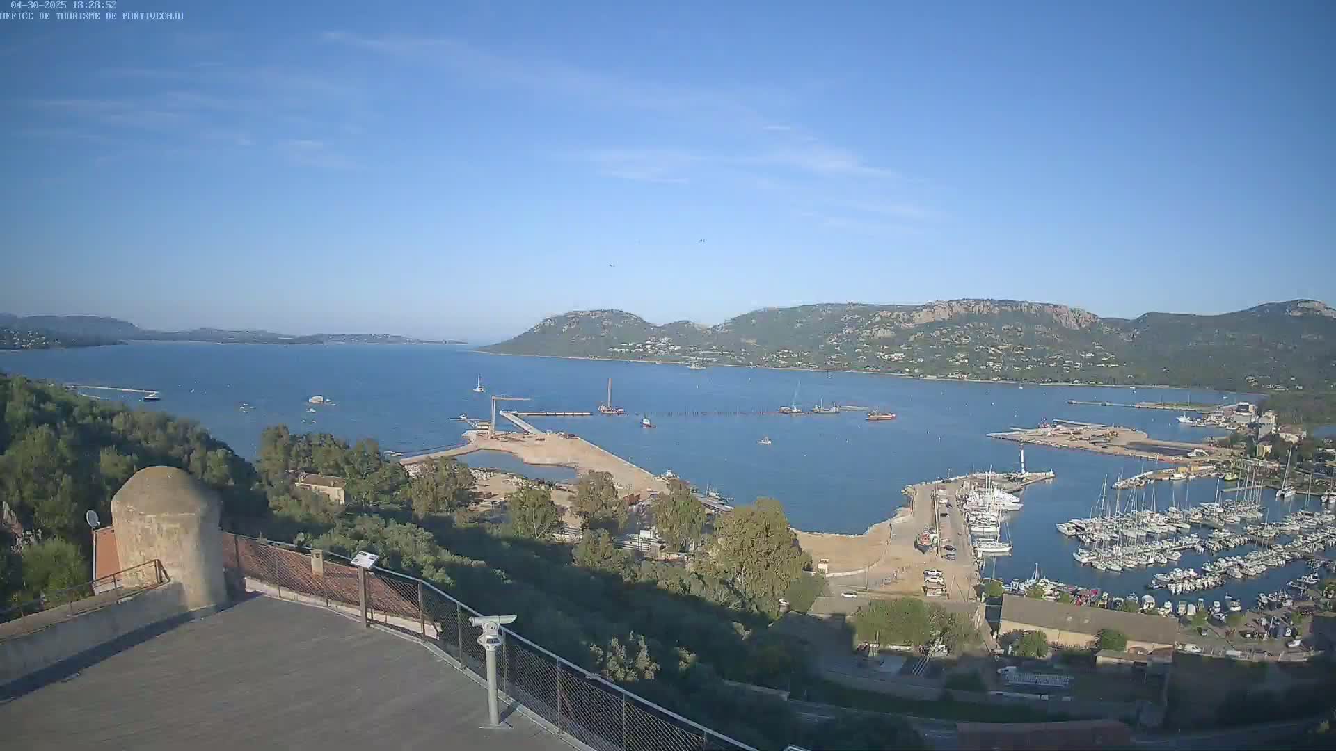 An elevated view of a calm bay on a sunny day shows a marina filled with sailboats, construction activity on a pier, and hills in the distance.