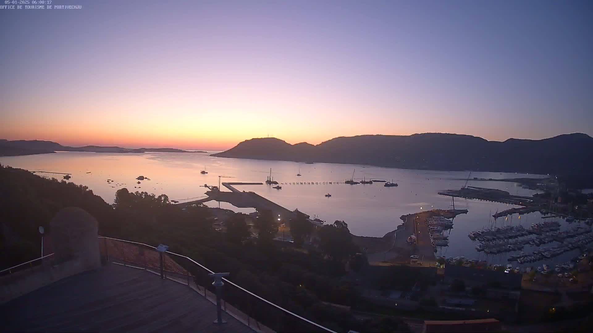 A calm harbor filled with boats is nestled between hills at sunrise under a clear, twilight sky.