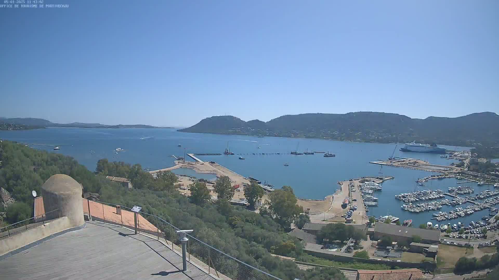 A high-angle view on a clear, sunny day shows a harbor filled with boats, a larger ship in the distance, and hills surrounding a body of water.