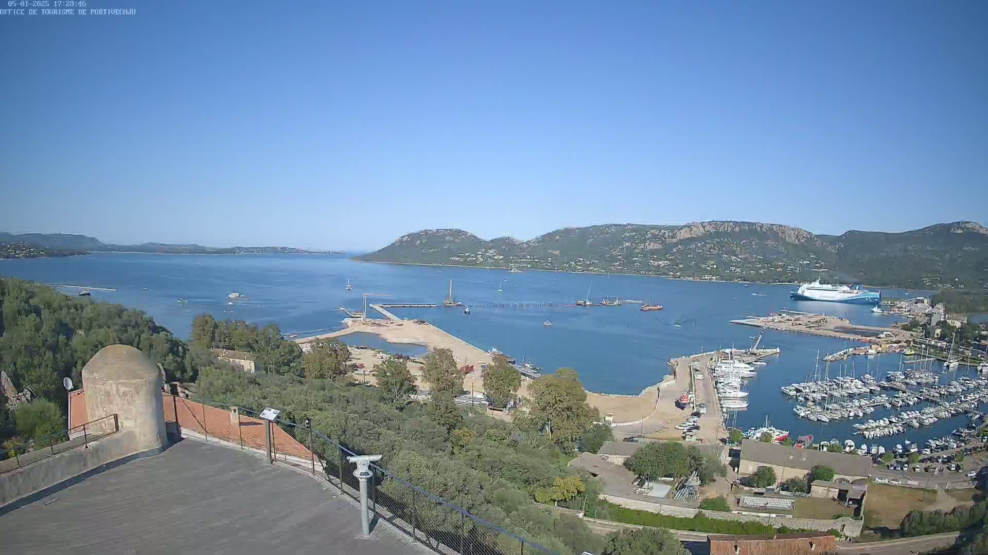 A high-angle, sunny view shows a harbor filled with boats and a large ferry docked near a sandy shoreline, with hills and calm blue water in the background.