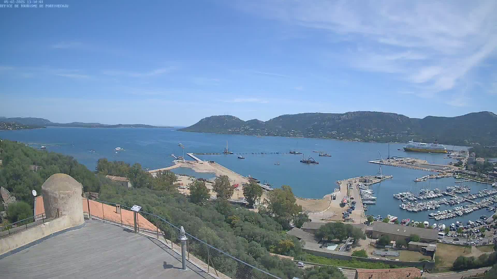 A sunny day overlooking a harbor filled with boats, a large ferry, and construction activity, all nestled between green hills and a calm bay.