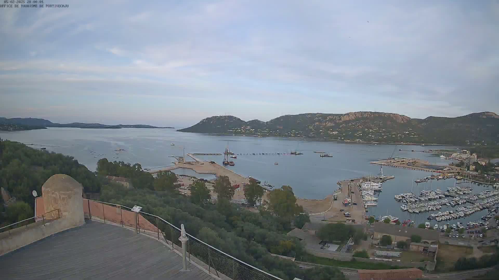 An elevated view of a calm bay with numerous boats docked in a marina, surrounded by hills under a mostly clear sky.